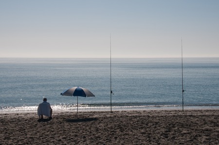 Lone surf fisherman sitting in a chair next to an sunshade at a beautiful beach on a sunny day doing fishingの写真素材