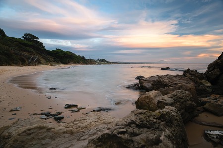 Landscape of a beach at the Costa de la Luz in Tarifa, Spain with a wooden stair as the entry to the beach. Photo taken at a colorful sunset, rocks in the foreground, Morocco in the background.の写真素材