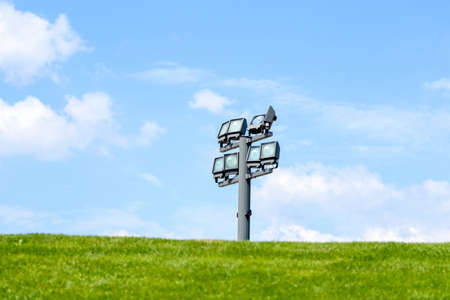 Electric spotlight on a metal pole during a sunny day on a green lawn. White clouds in the blue sky in the backgroundの写真素材