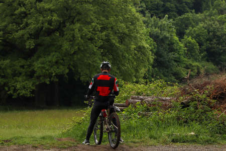 Man in red-black clothes on a bicycle in the woods. Thinking person on a mountain bike in the park. Trees in the background.の写真素材