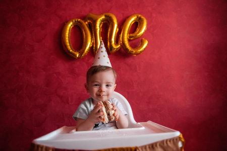 Little boy of one year old in a party hat sits in a children's chair, eating a birthday cake with a candle. Kid on a background of a red isolated background, on a textured wall foil gold balloons Oneの写真素材