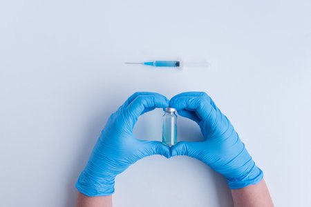 On white isolated background, the doctors hands in medical blue gloves depict heart shape gesture. Doctor is holding an ampoule with vaccine against virus. Blurred syringe with medicine. Copy spaceの写真素材