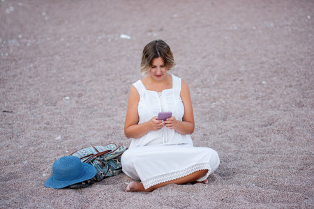 Middle age pregnant woman in white sundress sits in lotus position on the sand by the sea, talking on smartphone. Traveler with backpack, hat. Lifestyle pregnancy tourism concept. Social mediaの写真素材