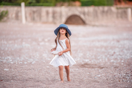 Little girl in blue hat, white dress plays on the sand, small pebbles on the seashore. Travel, holidays with children. Child stands tower of stones by the sea. Summer childhood. Copy spaceの写真素材