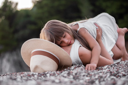 Millennial mother with daughter fooling around, having fun on the sand of the beach at sea. Little girl hugs middle-aged woman in hat, tickles mom. The journey of single parent. Travel trip pregnancyの写真素材