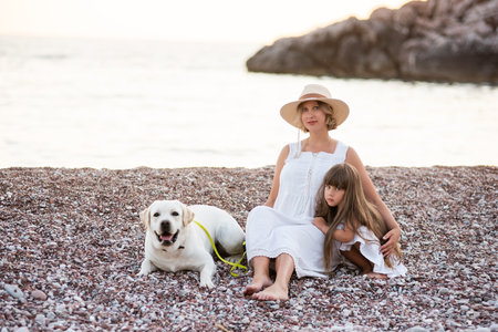 Pregnant mother with little daughter are sitting along the sand of the beach near the sea with golden retriever. Millennial woman holds dog on leash, girl by the hand. Concept of traveling with petsの写真素材