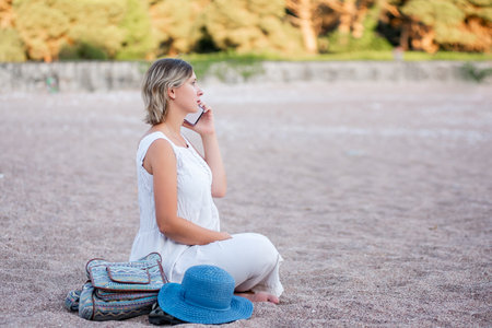 Middle age pregnant woman in white sundress sits in lotus position on the sand by the sea, talking on smartphone. Traveler with backpack, hat. Lifestyle pregnancy tourism concept. Social mediaの写真素材