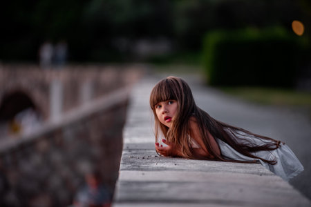 Little girl in white dress with long hair looks at the sea, stands on the parapet, looks at the mountains in the sunset light. Bright childhood filled with new discoveries. Family travel with childrenの写真素材