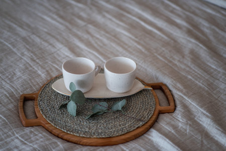On the Linen bedspread lies a wooden round tray, on it are two empty ceramic cups, green eucalyptus. Stylish, trendy morning. Textural, Scandinavian Rustic interior. Light gray background. Copy spaceの写真素材