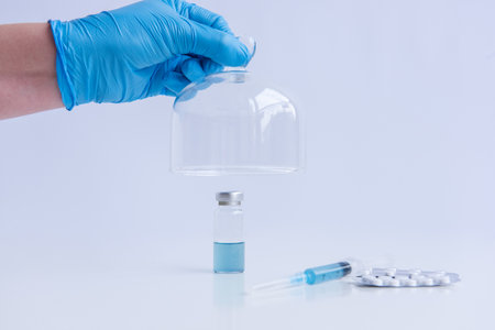 On white isolated background stands the vaccine with syringe, pills. Doctors hand in blue medical glove opens programmed protective dome. Cure against viruses, treatment of people. Mock up, copy spaceの写真素材