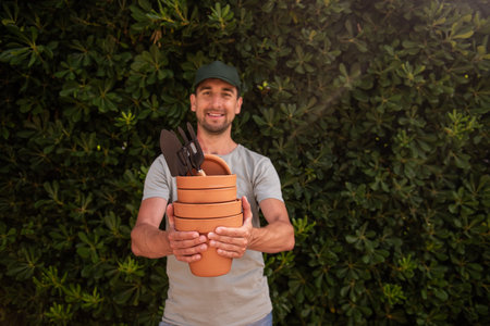 Male gardener in green cap holds terracotta clay pots with gardening supplies in hands. Looks at the camera, smiles. Middle-aged man with red birthmark on neck in front of an evergreen fence. Mock upの写真素材