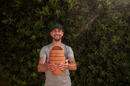 Close-up portrait man gardener in green cap holds terracotta clay pots in hands against the backdrop of an evergreen fence. Gardening, small business, hobby. Trendy garden landscaping. Copy spaceの写真素材