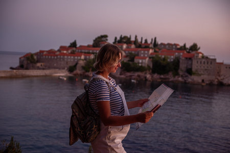 Middle aged pregnant woman in vest holds map, phone in hands. Traveler with backpack looks at the island of Sveti Stefan at sunset in Montenegro. Travel motherhood pregnancy concept. Summer vibesの写真素材
