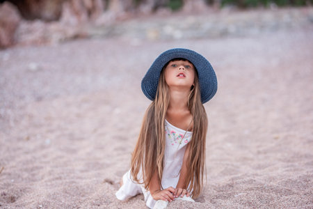 Little girl in blue hat, white dress plays on the sand, small pebbles on the seashore. Travel, holidays with children. Child stands tower of stones by the sea. Summer childhood. Copy spaceの写真素材