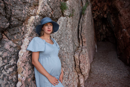 Pregnant millennial woman in blue dress stands among the rocks on the seashore, hugging belly with her hands. Girl in hat on background of textured wall. Natural beauty concept. Copy space, travellerの写真素材