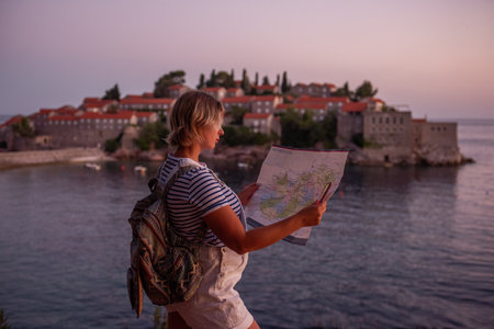Middle aged pregnant woman in vest holds map, phone in hands. Traveler with backpack looks at the island of Sveti Stefan at sunset in Montenegro. Travel motherhood pregnancy concept. Summer vibesの写真素材