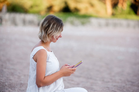 ?lose-up portrait of a woman in a white sundress sits on the sand by the sea, typing a message on a smartphone. Green phone screen mockup. Internet connection in tourism. Faceless, copy space, mock upの写真素材