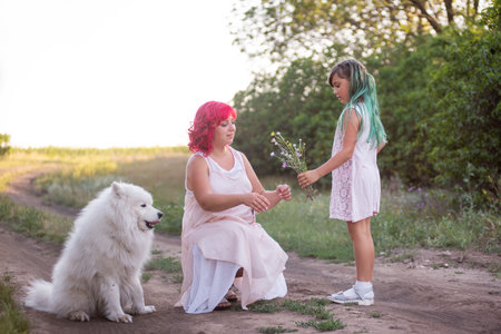 Girl with green hair gives wild flowers to mother with pink. Traveling with Samoyed dog, pets to nature. The tenderness of motherhood, one parent. Diversity family. Beautiful paths in the forest.の写真素材
