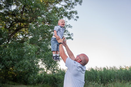 Bald man with glasses throws child into the sky air. Father in jeans plays, embrace with son in nature outside the city. Little boy laughs, having fun with dad on journey in the forest. Fathers dayの写真素材