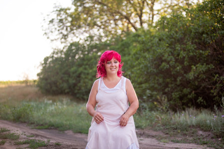 Bright diversity woman with pink hair portrait. The girl squatted down on a forest path into the sunset. Childfree girl, weekend trip. Individual, bold style. Green nature background. Copy spaceの写真素材