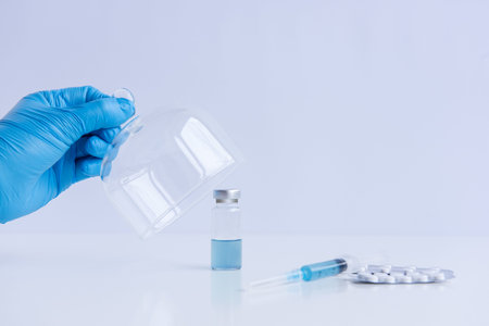 On white isolated background stands the vaccine with syringe, pills. Doctors hand in blue medical...