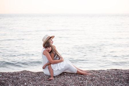 Little girl sits in the arms of pregnant mother on the sand by the sea on beach. Daughter hugging millennial woman in hat. The journey of single parent. Travel pregnancy, copy space. Summer vibesの写真素材