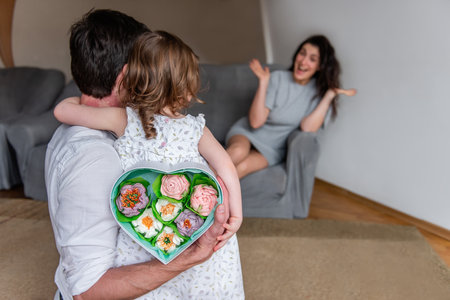 Little daughter and father hold gift for mother behind backs, heart shaped box with homemade marshmallow flowers. Surprise for woman on mothers day from child and husband. Girl is emotionally happyの写真素材