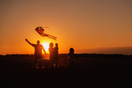 Panorama silhouette diversity family with Samoyed running at sunset with kite in sky. View from below through the ears. Father holding toy in hands, mother embrace little son, daughter with dogの写真素材