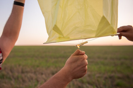 Mans hand sets fire to heavenly yellow sky lantern with lighter. Making wishes for the holidays. The concept of hope, faith. Paper, Chinese lantern. Teamwork. Copy space. Facelessの写真素材