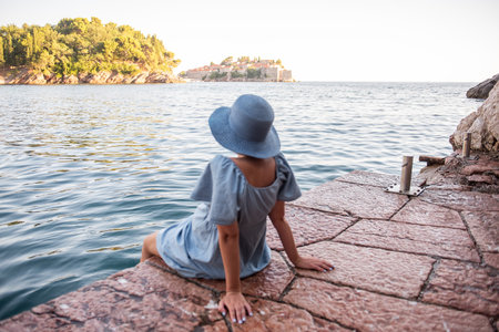 Defocesed Faceless Pregnant woman traveler in denim dress, blue straw hat sits by sea, Looks at island of Sveti Stefan in Montenegro at sunset. Tourism, travel in pregnancy. Healthy lifestyle. Summerの写真素材