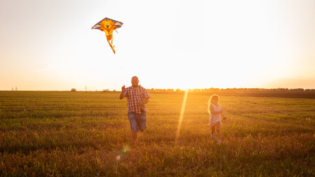 Bald father in glasses runs around the field with daughter putting into the sky kite. Millennial man plays with girl in rural areas. Traveling, fun on the day of the father. Warm happy family memoriesの写真素材