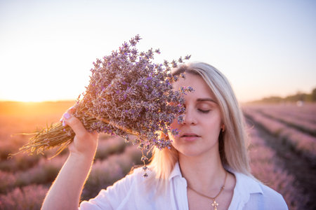 Close-up portrait of young woman with bouquet of purple lavender in front of face. Blonde millennial girl with clean skin without makeup in rays of sunset. Allergy concept. Aroma oils natural perfumesの写真素材