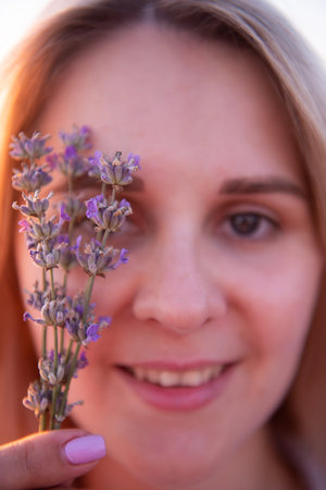 Sprigs purple lavender against background of blurred face of young blonde woman. Close-up portrait of millennial girl with clean skin without makeup. Allergy concept. Aroma oils, perfumery from natureの写真素材