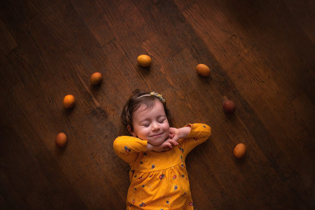Little girl lies on brown wooden floor in dress among Easter yellow eggs painted with natural...