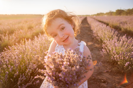 Close-up portrait of little girl in flower dress holding bouquet with purple lavender at sunset. Child stands among the rows in field. Walk in countryside. Allergy concept. Natural products, perfumeryの写真素材