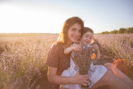 Portrait of brunette mother with little daughter sitting in purple lavender field. Young woman in rural dress lovingly embraces, kisses girl. The concept of allergy, travel, single parent. Copy spaceの写真素材