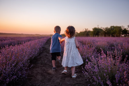 Playful cute boy girl are playing in rows of lavender purple field at sunset. Small couple runs after each other, catches up, holding hands. Cheerful, happy childhood. Travel in countryside. Allergyの写真素材