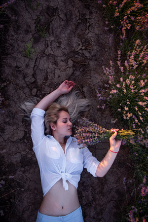 Young blonde woman lies on the ground between rows of purple field at sunset. Childfree romantic girl in white shirt holds bouquet in hands. Travel in the countryside. Concept of allergy, freedomの写真素材