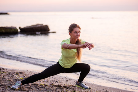Young woman in sportswear does sit-ups early in morning on seashore at sunrise. Girl of slim build with long hair is stretching. Outdoor sports by the water. Fitness classes, body care, mental healthの写真素材