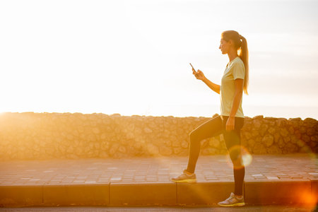Close-up portrait of young woman in sportswear, holding smartphone, takes selfie in between workouts against the backdrop of an orange dawn sky. The concept of sports and communications. Cyberspaceの写真素材
