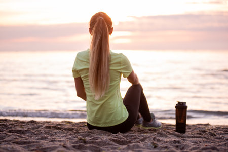 Young woman in sportswear is sitting on the seashore at sunrise. Girl is resting after workout, replenishes her water balance, drinks from a sports bottle, looks at the water. Morning outdoor fitness classes.の写真素材