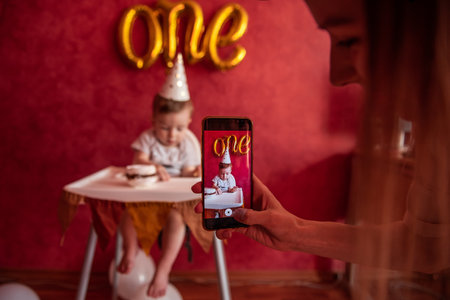 Mothers hand takes pictures of son on a smartphone. A little boy of one year in festive hat holding birthday cake with a candle in hands. On red plain background gold foil balloons One. Social Mediaの写真素材