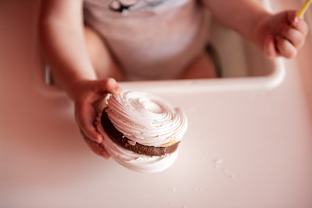 A faceless portrait of a toddler sitting in a feeding chair holding a mini meringue cake with chocolate. Homemade sweets. Useful dessert. Birthday cake for a one year old. Tactile foodの写真素材