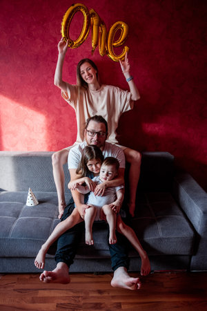 Portrait of happy, diversity family on the birthday of one year old boy. Mother laughs, holds foil balloons ONE in hands, father hugs daughter and son on gray sofa in the sun. Home holiday party.の写真素材