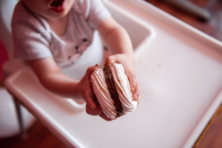 A faceless portrait of a toddler sitting in a feeding chair holding a mini meringue cake with chocolate. Homemade sweets. Useful dessert. Birthday cake for a one year old. Tactile foodの写真素材