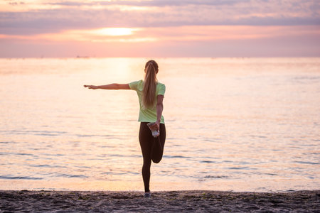 Young woman in sportswear is warming up early in morning on seashore at sunrise. Girl of slim build with long hair is stretching. Outdoor sports by the water. Fitness classes, body care, mental healthの写真素材