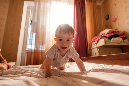 One year old baby lies on his stomach on the bed in the morning in the sun. The child is funny,...