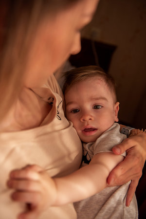 Young, stylish blonde mother holds little son in arms near the window. Caring for the baby,...