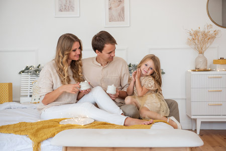 Funny mother father are drinking cocoa with marshmallows from white cups on bed, looking at cute daughter. Christmas interior, cozy holiday. Young family hugs, laughs. Curly girl looks at the cameraの写真素材