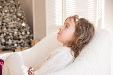 A pensive, dreamy little girl lies in a soft white armchair, dreams of a gift from Santa Claus near the Christmas tree. Child in a cozy atmosphere at home. Festive interior. Copy spaceの写真素材